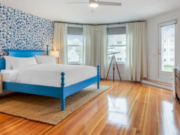 A bedroom with a blue bed, patterned wall, wood floor, dresser, and two windows with curtains. Natural light fills the space from large windows.