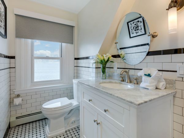 A bright bathroom with white tiles, featuring a circular mirror, sink with flowers, and a toilet. A window brings in natural light.