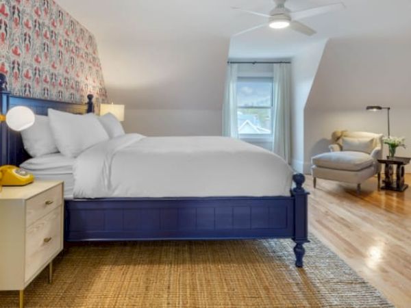 A cozy bedroom with a blue bed, patterned wallpaper, white nightstand, and wooden floor. A chair, lamp, and window add to the decor.