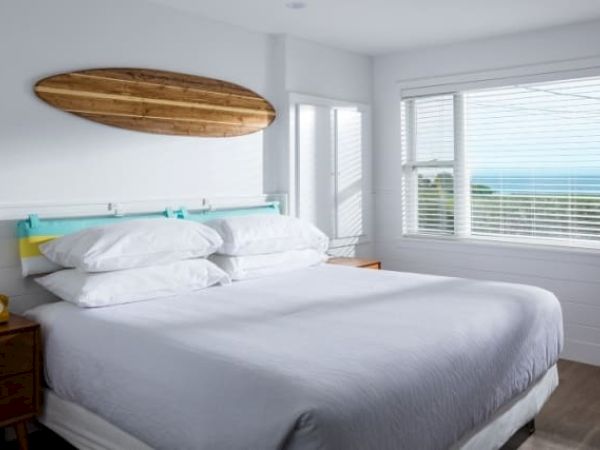 A bedroom with a white bed, wooden surfboard decor, large windows, and a view of the ocean under clear blue skies.