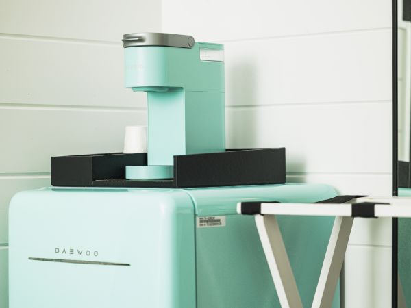The image shows a mint green coffee machine on a matching fridge, alongside a white and black folding table in a white-tiled room.