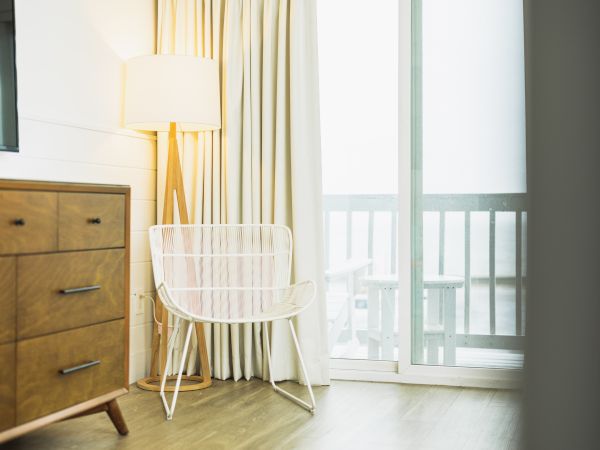 A cozy room with a wooden dresser, white chair, and tall lamp beside a curtained sliding glass door opening to a balcony.