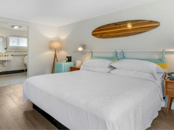 A cozy bedroom featuring a white bed, modern nightstands, a surfboard decor, and a lit lamp, with a glimpse of the bathroom.