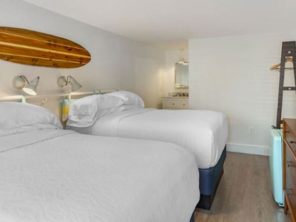 A hotel room with two beds, surfboard wall decor, desk with TV, and a ladder holding clothes hangers, in a clean, contemporary style.