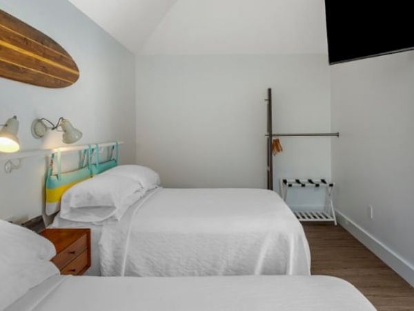 The image shows a bedroom with two single beds, a surfboard decoration, overhead lights, and a flat-screen TV on the wall.