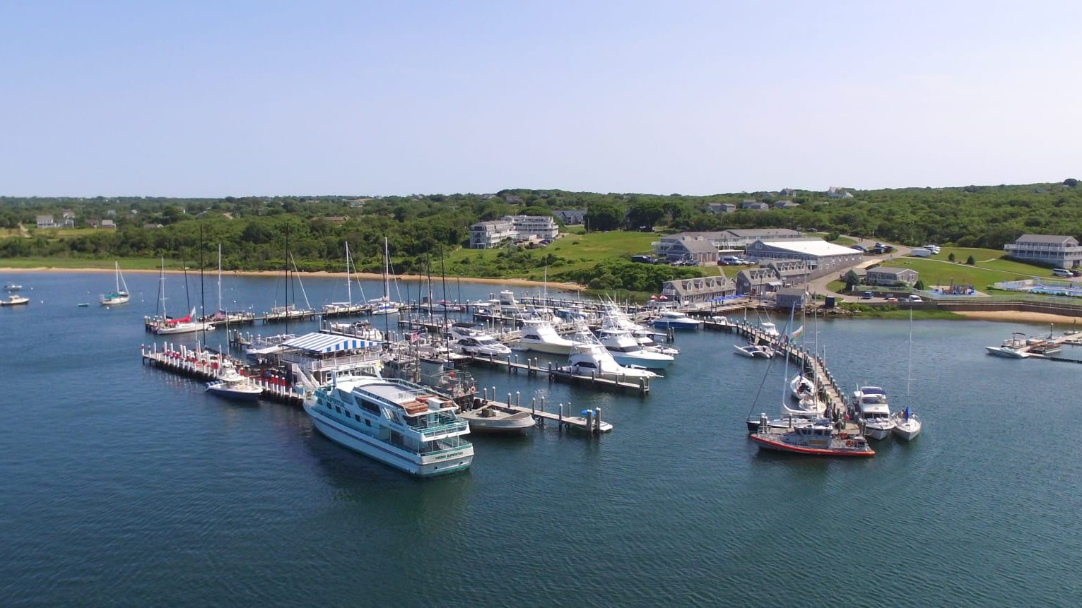The image shows a marina with multiple boats and yachts docked, surrounded by calm water and some buildings in the background.