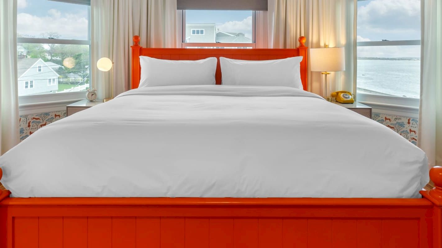 A bedroom with an orange wooden bed, white bedding, curtains, bedside lamps, and a view of the sea or buildings through the windows.