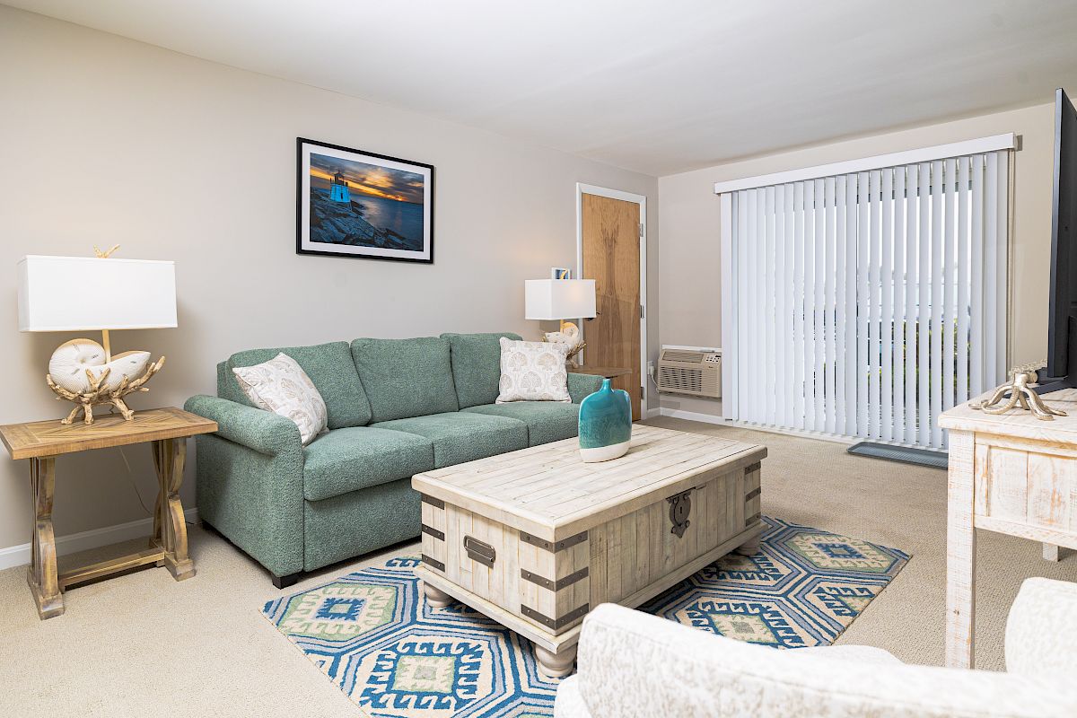 A cozy living room with a green sofa, wooden coffee table, lamps, decorative art, and a large window with blinds.