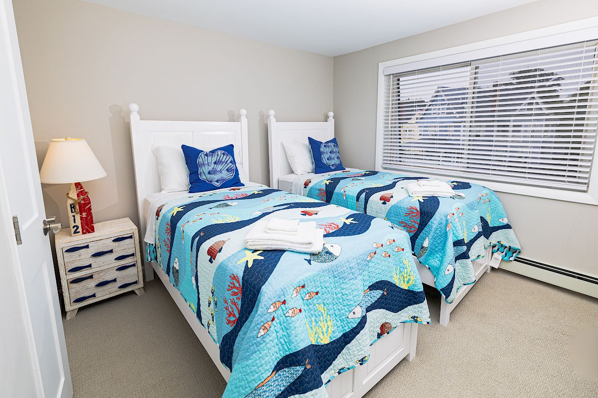 The image shows a bedroom with two twin beds, each with blue ocean-themed bedding, a lamp, and a small dresser by the window.