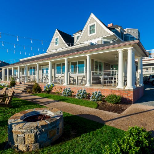 A large house with a porch, Adirondack chairs, fire pit, string lights, and blue skies, creating a cozy outdoor atmosphere.