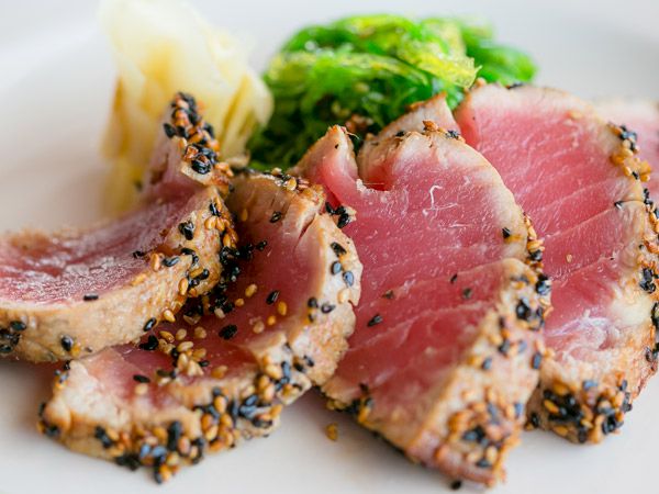 Sliced sesame-crusted seared tuna with a pink center, served on a white plate with greens and light sauces, plated elegantly.