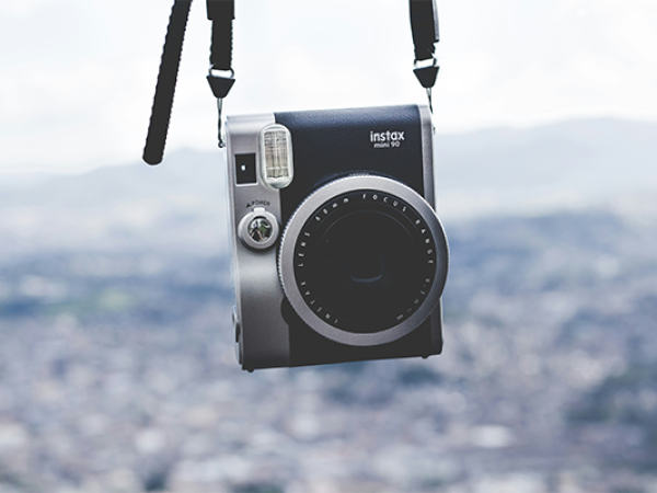 A black and silver instant camera hangs by its strap against a blurred cityscape background.