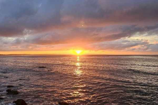 A stunning sunset over the ocean with a rocky shoreline and dramatic clouds in the sky.