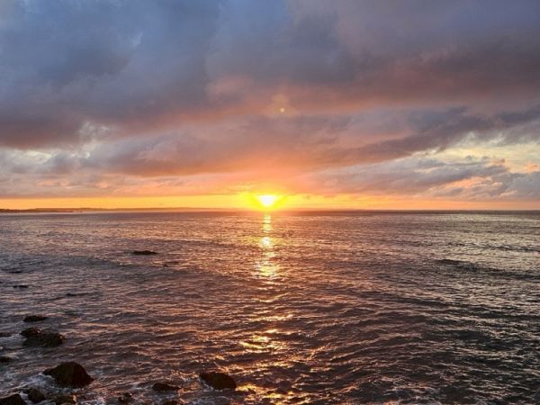 A stunning sunset over the ocean with a rocky shoreline and dramatic clouds in the sky.