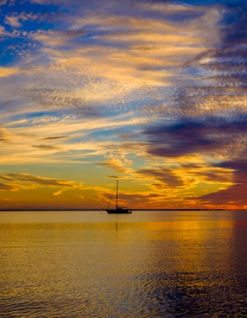 A serene sunset over calm waters with a silhouetted sailboat in the distance and vibrant, colorful clouds in the sky.