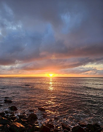 A stunning sunset over the ocean with clouds, reflecting vibrant colors on the water's surface and rocky shoreline.
