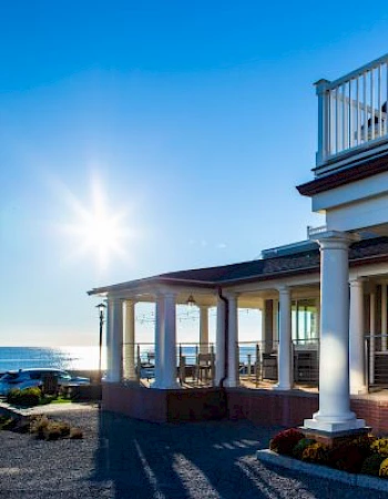 A sunny coastal house with a wraparound porch and white columns, bright blue sky, and ocean view, golden hour lighting.