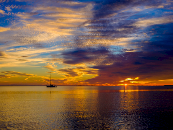A serene sunset over calm waters, with a silhouetted sailboat in the distance and dramatic clouds and vibrant colors filling the sky.