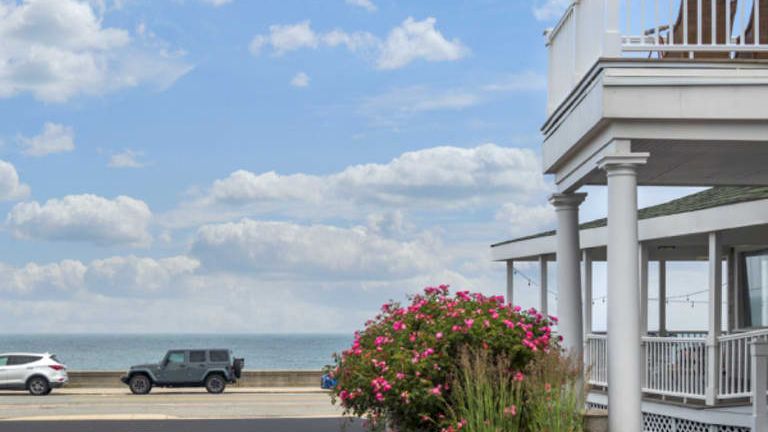 A seaside scene with a two-story porch building to the right, pink flowering shrub, parked cars along the road, and a sunny sky over the ocean.