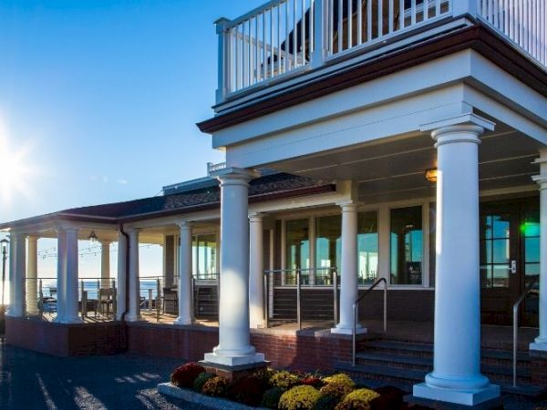 A seaside house with white columns and a covered porch, sunshine sparkling off the ocean, balconies above, cozy exterior lighting.