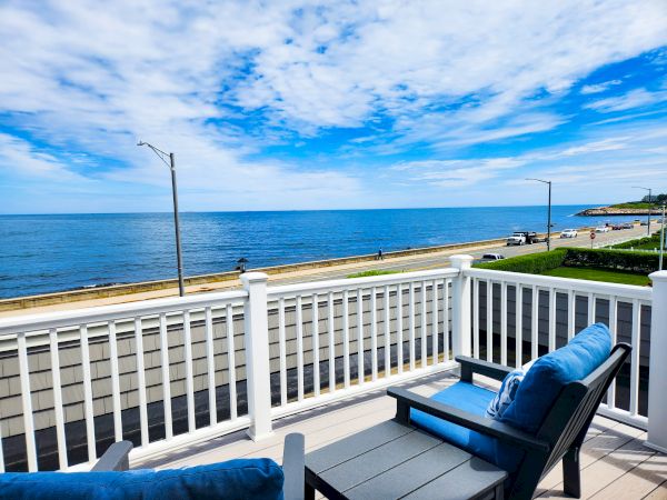 A sunny seaside view from a balcony overlooking the ocean, with blue chairs and white railings, a clear sky, and a quiet coastal road.