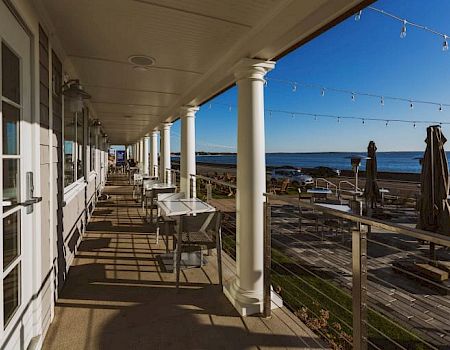 A seaside porch with white columns, tables and chairs, sunlit deck, string lights, and a view of the beach and ocean in the distance.