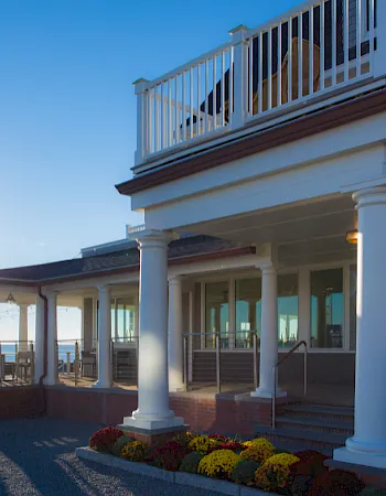 A sunny coastal restaurant with white columns, a wraparound deck, and colorful flowers near the entrance; sunlit and serene.