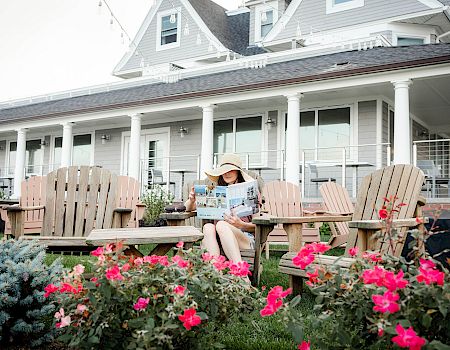 A person sits on a wooden chair on a porch, reading a magazine, with pink flowers in front and a large house in the background.