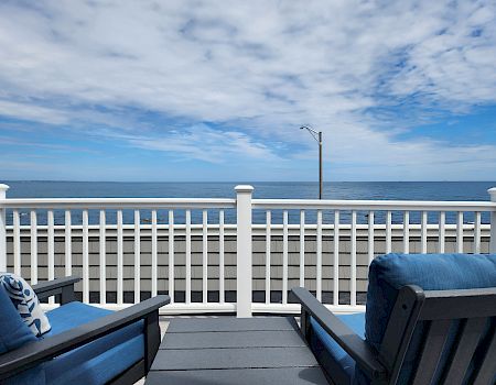A seaside porch with two blue cushioned chairs facing the ocean, white railing, and a clear sky above the calm water.