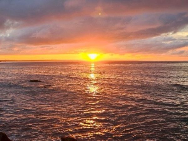 A stunning sunset over calm waves, with a glowing orange sun dipping toward the horizon and warm pinks lighting the cloudy sky.