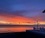 Sunset over the ocean with vivid orange and purple skies, framed by a seaside deck railing and lamp lights.