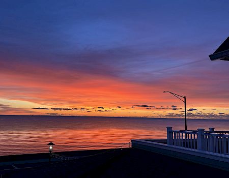 Sunset over the ocean with vivid orange and purple skies, framed by a seaside deck railing and lamp lights.