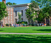 A sunny college campus scene with students walking on the green lawn beside brick and gray academic buildings.