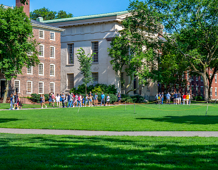 A sunny college campus scene with students walking on the green lawn beside brick and gray academic buildings.