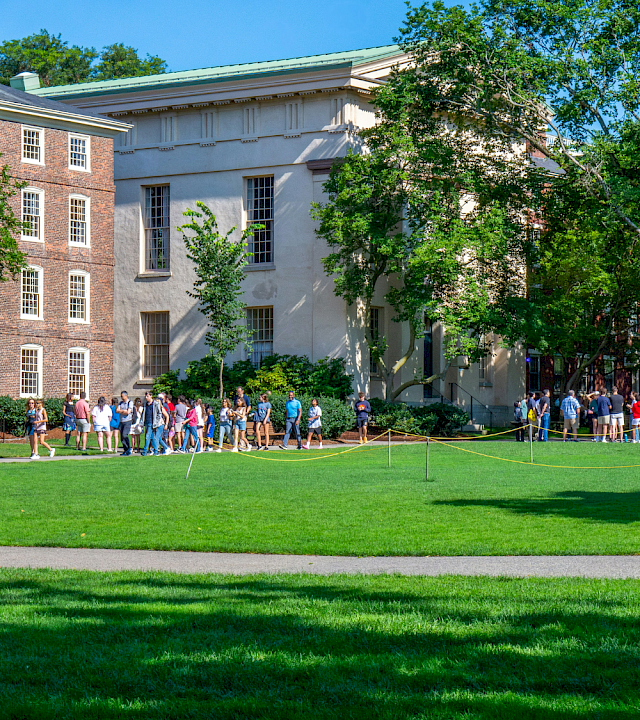 A sunny college campus scene with students walking on the green lawn beside brick and gray academic buildings.