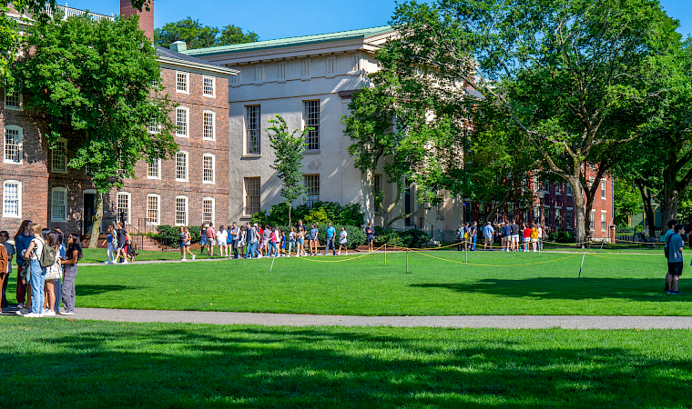A sunny college campus scene with students walking on the green lawn beside brick and gray academic buildings.