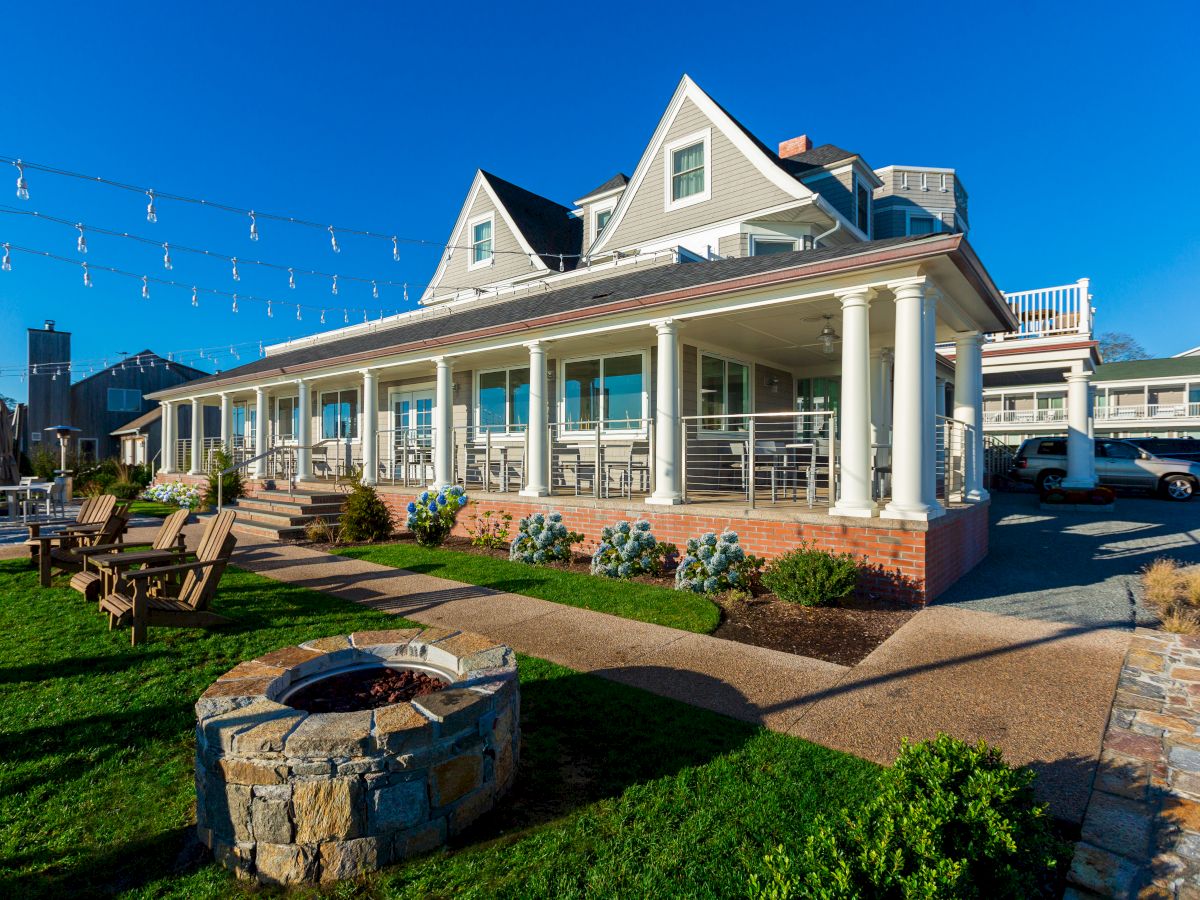 A sunny view of a spacious house with a covered front porch, white columns, string lights, flower beds, and a driveway.
