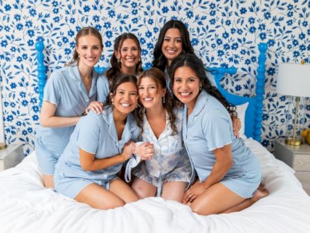 A group of women in blue scrubs are smiling together on a bed in front of blue floral wallpaper, posing for a photo.