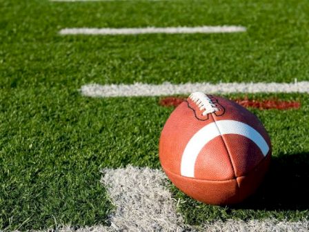 An American football rests on green grass turf near the white yard line, with a few wisps of artificial turf around it.