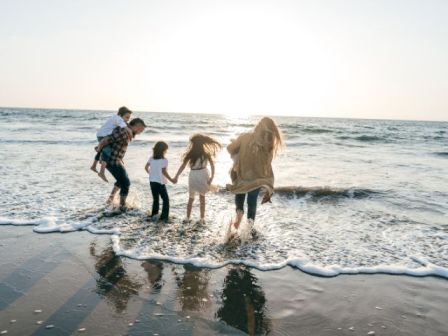 A family of four runs and plays in shallow ocean waves at sunset, laughing on the beach.