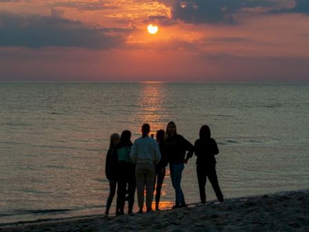 A group of friends stands on a beach watching the sun set over the ocean, casting golden reflections on the water.