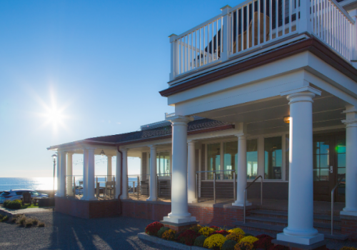 A seaside porch with white columns on a blue-sky morning, bright sun and colorful flowers in front.