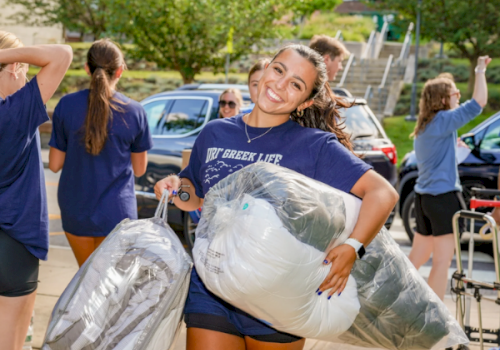 A smiling volunteer in a blue T‑shirt carries large bags of donated items while others sort and stretch outdoors.