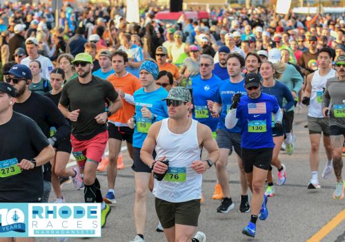 A packed street race with hundreds of runners in numbered bibs, jogging toward the camera under city lights.
