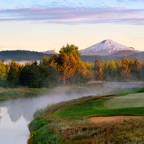 A golf course with a river, lush trees, and distant mountains under a clear sky at dawn.